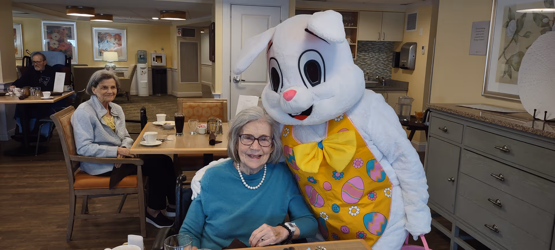 Elderly residents in a senior living dining room, one smiling while posing with a person dressed as an Easter bunny.