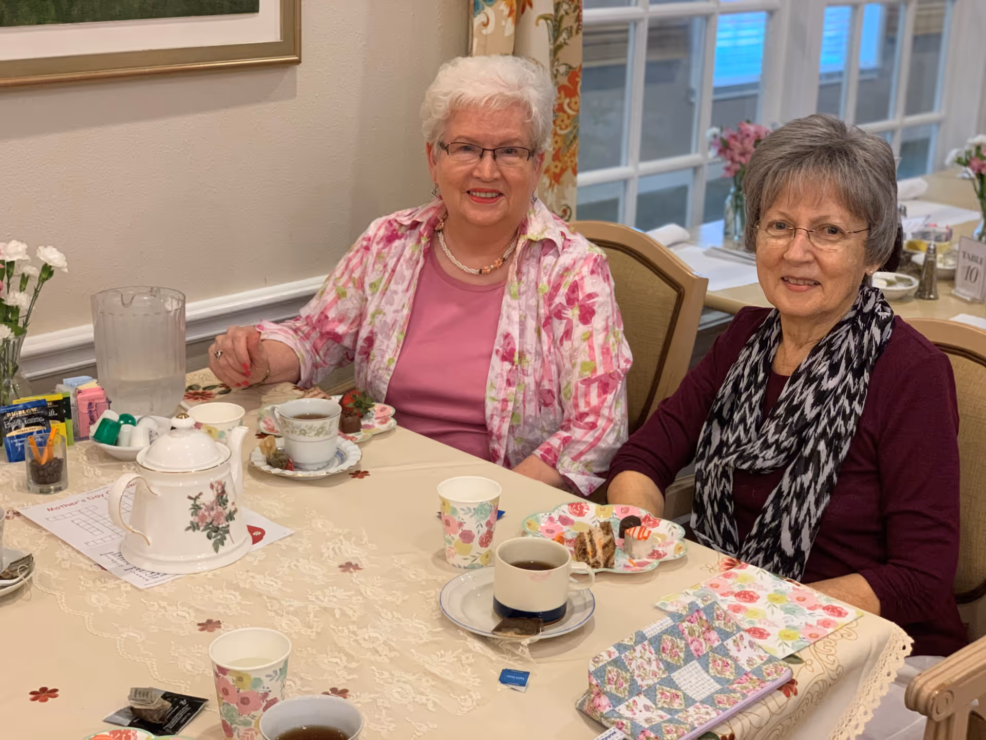 Two elderly women sitting at a dining table with tea cups, plates of desserts, a teapot, and a pitcher of water. The table is covered with a lace tablecloth and decorated with floral napkins and a small vase of white flowers. The background shows a window with floral curtains and additional tables set for dining.