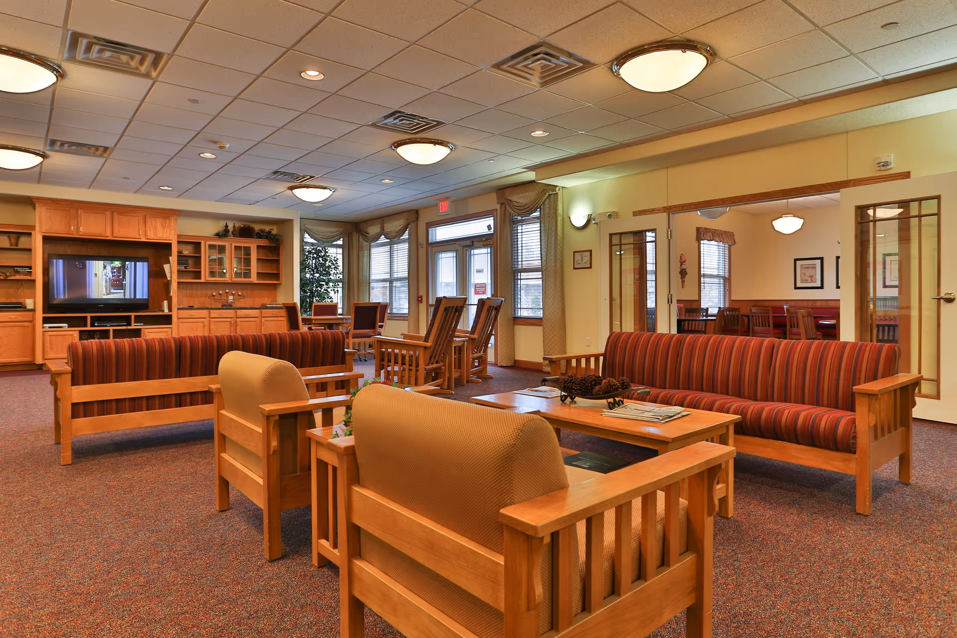 A spacious senior living facility common area with wooden furniture including striped sofas, armchairs, and coffee tables. The room has a carpeted floor, ceiling lights, and large windows with curtains allowing natural light. There is a built-in wooden entertainment center with a TV, and a dining area is visible through glass doors on the right.