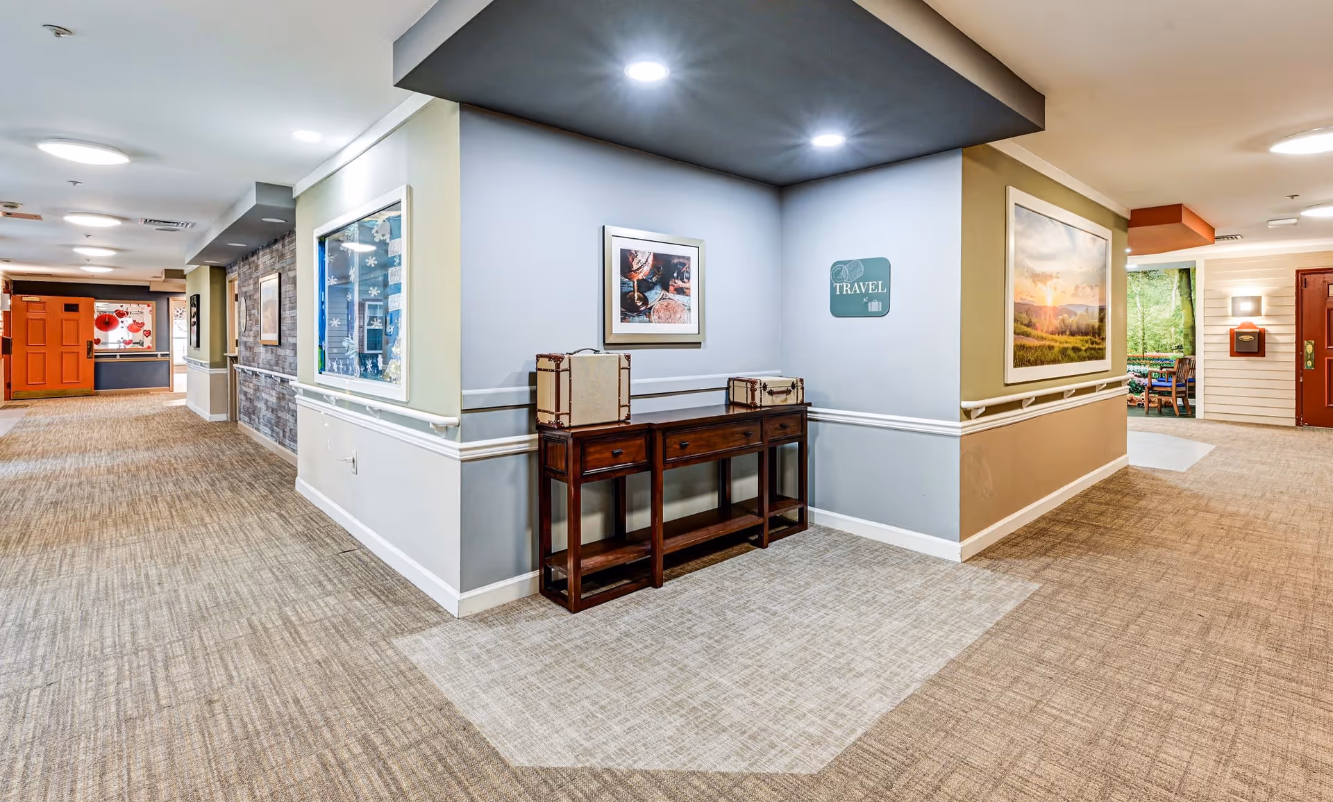A well-lit interior hallway in a senior living facility with beige carpet and light-colored walls. The hallway features a wooden console table with decorative suitcases on top, framed artwork on the walls, and handrails along the walls. The ceiling has recessed lighting and a dark accent area above the console table. The hallway extends in two directions with doors and additional artwork visible.
