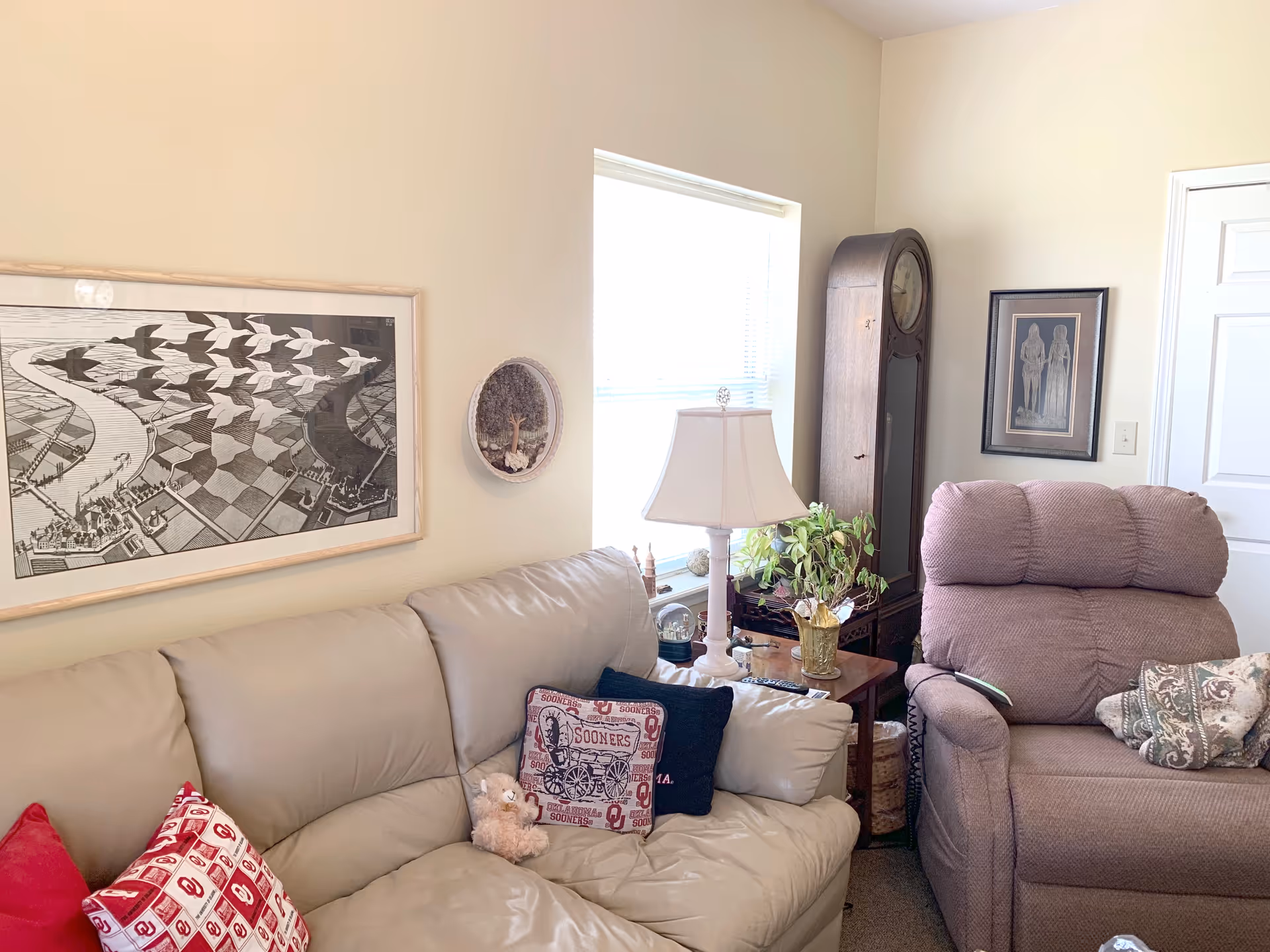 A cozy living room with a beige leather sofa adorned with decorative pillows and a small teddy bear. Next to the sofa is a wooden side table with a white lamp and a potted plant. A tall grandfather clock stands in the corner beside a framed picture on the wall. The room has light-colored walls and a window letting in natural light.