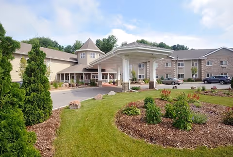 Exterior view of a senior living facility with a covered entrance, landscaped garden with shrubs and flowers, and a parking area with several cars. The building has multiple windows and a turret-like architectural feature.