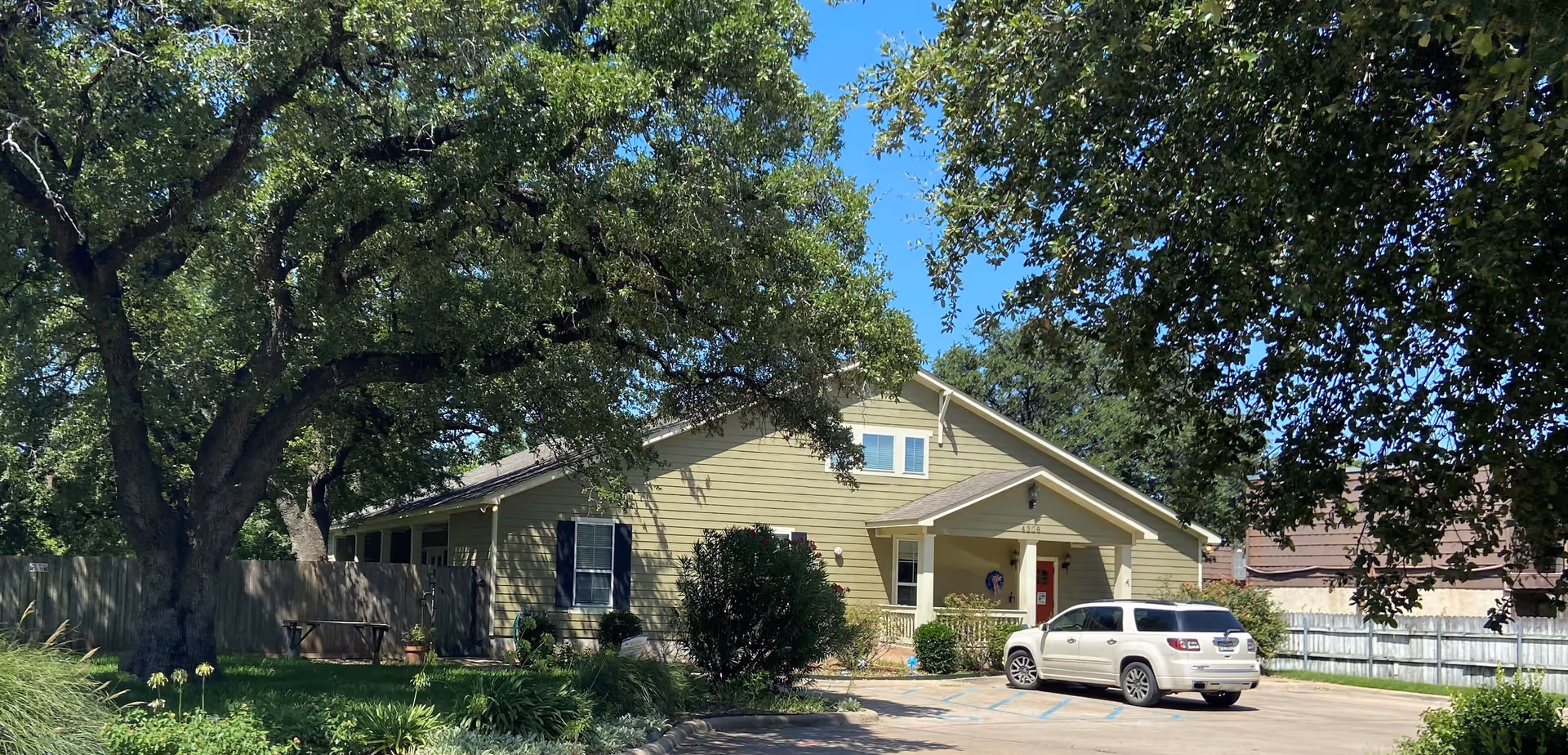 Exterior view of a single-story building with light green siding and a small covered porch. A white SUV is parked in front on a paved driveway. Large trees with green foliage surround the building, and there is a wooden fence and garden area with plants and a bench to the left.