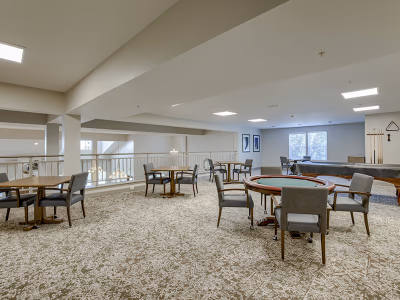 A spacious recreational room with multiple round tables and chairs arranged for group activities. The room features a carpeted floor, neutral-colored walls, and ceiling lights. In the background, there is a pool table with cues mounted on the wall and two windows letting in natural light.