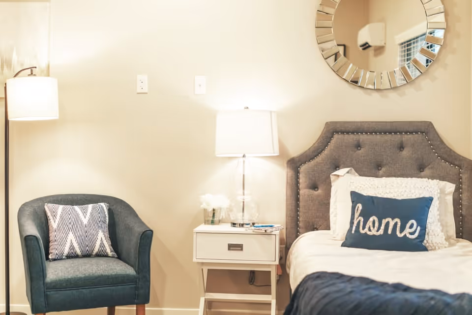 A cozy bedroom corner featuring a gray upholstered bed with a decorative 'home' pillow, a white nightstand with a clear glass lamp and small flower arrangement, a blue armchair with a patterned cushion, and a round decorative mirror on the beige wall.