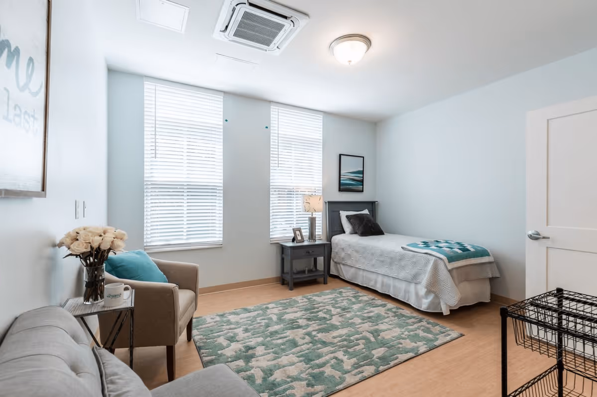 A bright and clean bedroom with light blue walls, two large windows with white blinds, a single bed with white bedding and a teal throw blanket, a gray nightstand with a lamp and picture frame, a beige armchair with a teal pillow, a small side table with a vase of white roses and a mug, a gray couch, and a patterned teal and white rug on a wooden floor.