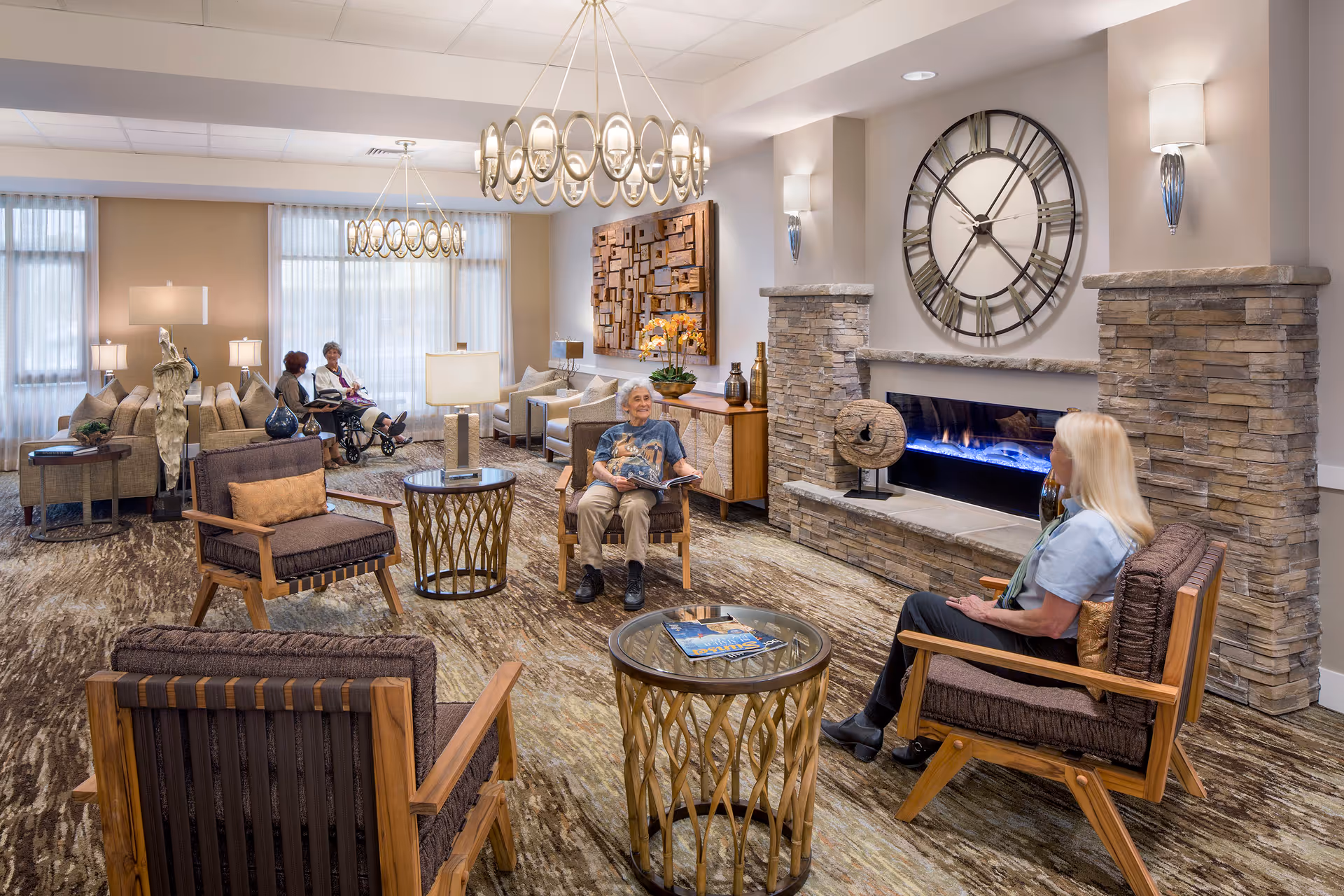 A cozy senior living facility lounge with comfortable seating, a modern stone fireplace with a large clock above it, and warm lighting. Several elderly residents are seated and engaged in conversation or reading. The room features large windows with sheer curtains, decorative wall art, and stylish round tables.