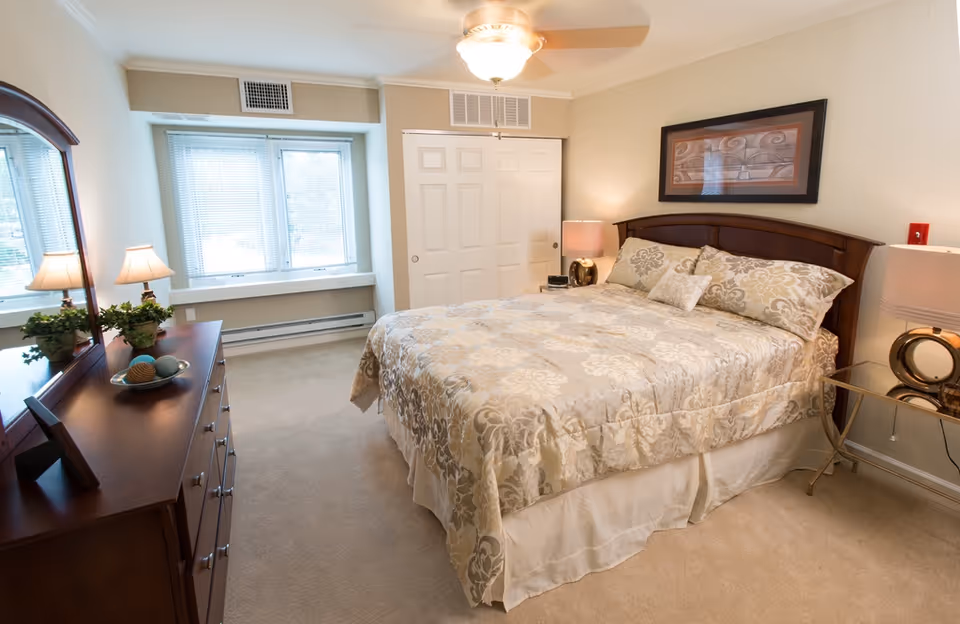A well-lit bedroom with a large bed covered in a beige and cream floral patterned comforter and matching pillows. There is a wooden dresser with a mirror on the left side, decorated with a small plant and decorative balls. Two bedside tables with lamps flank the bed. A window with blinds is on the far wall, and a closet with sliding doors is visible next to the window. The walls are painted light beige, and a framed artwork hangs above the bed.