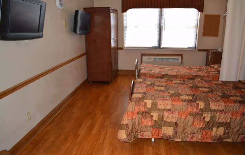 A bedroom with two single beds covered in patchwork bedspreads, wood flooring, a wall-mounted TV, armoire and a window.