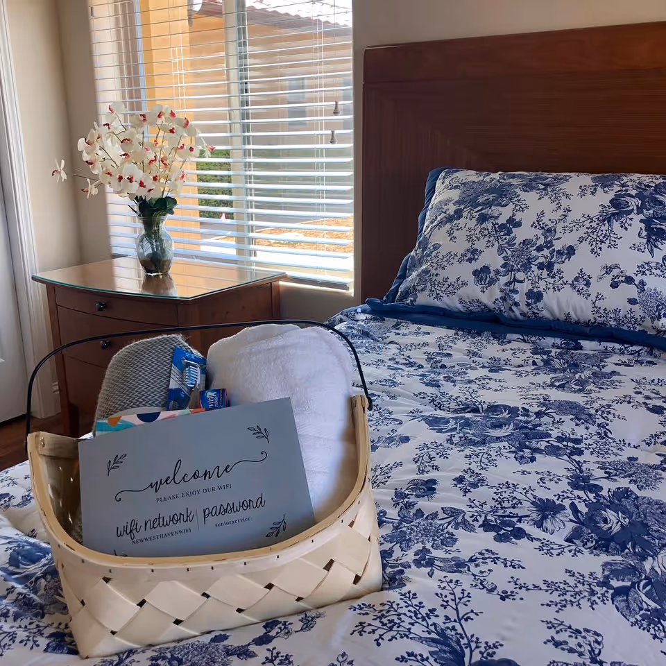 A neatly made bed with blue and white floral bedding in a bedroom. On the bed is a woven basket containing a white towel, a gray item, and a card with wifi network and password information. Next to the bed is a wooden nightstand with a glass vase holding white orchids. A window with blinds is in the background, letting in natural light.