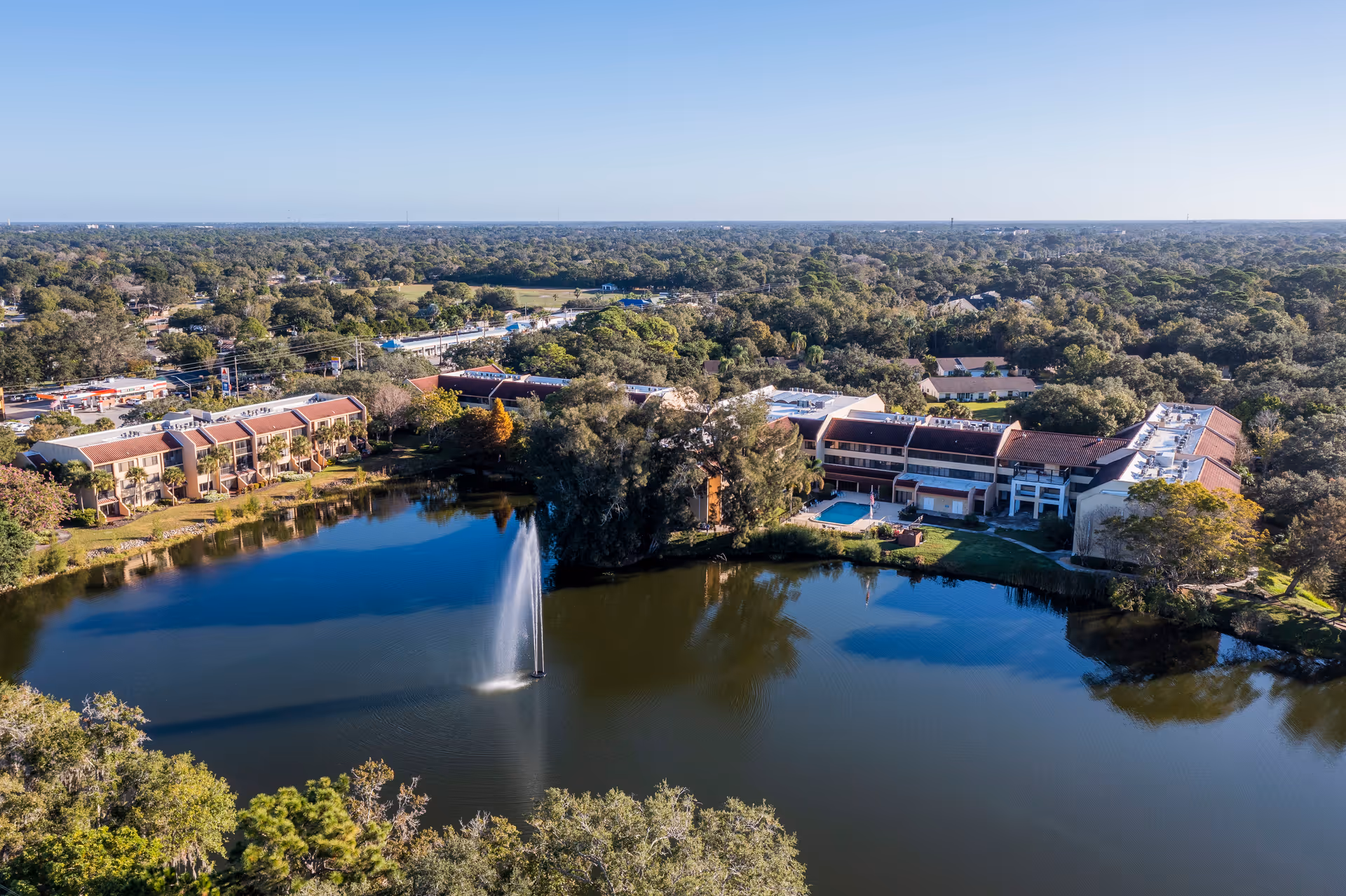 Aerial view of American House Sarasota senior living facility showing multiple buildings surrounding a large pond with a water fountain in the center, surrounded by trees and greenery under a clear blue sky.