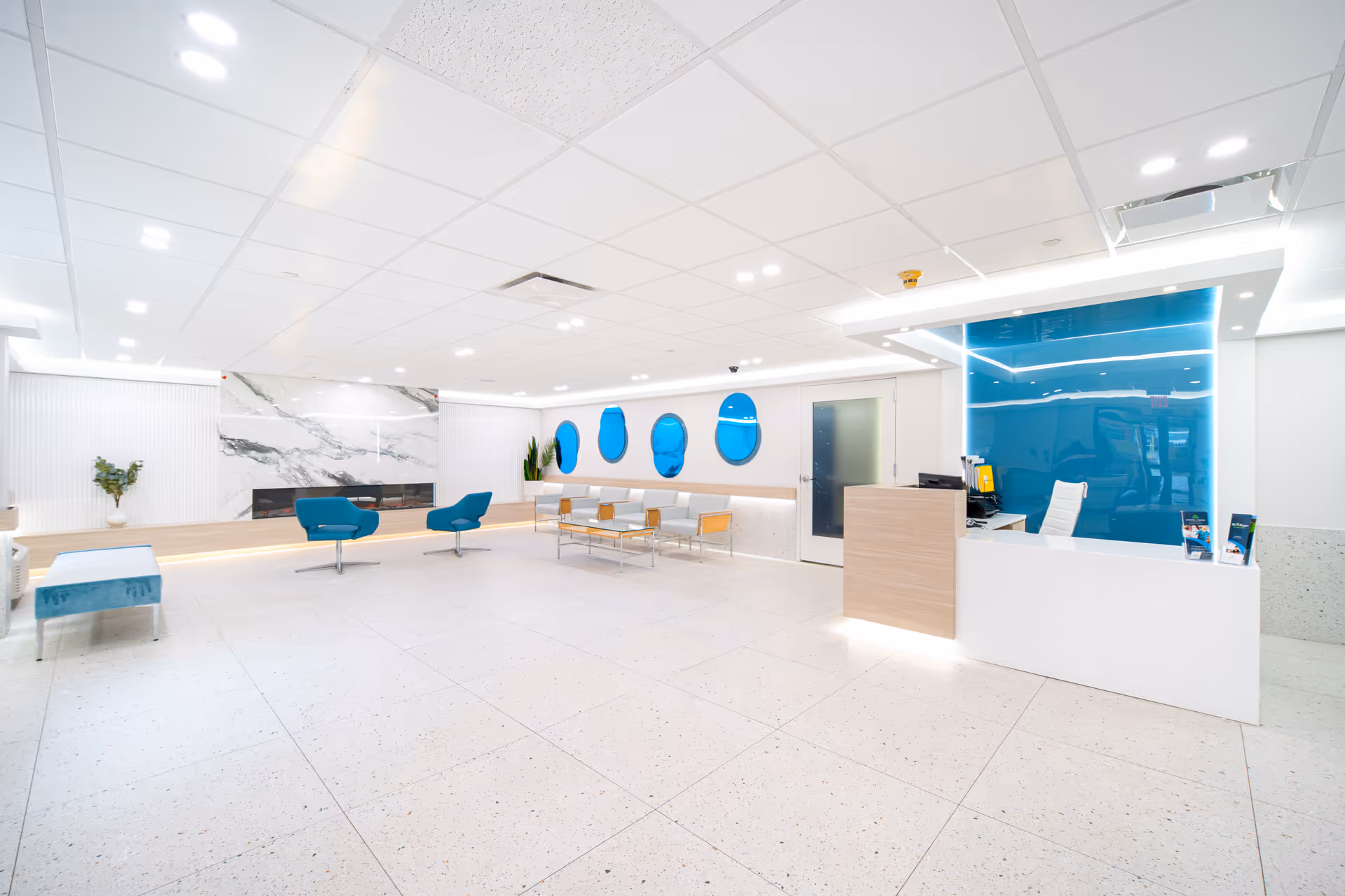 A spacious, modern healthcare center lobby with white tiled floors and ceiling. The reception desk is on the right with a blue glass panel behind it. There are blue chairs and a bench seating area with a glass coffee table in the center. The back wall features a marble design and blue oval windows. The space is brightly lit with recessed ceiling lights.