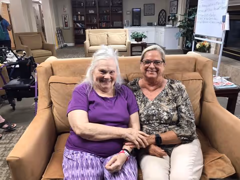 Two elderly women sitting closely together on a tan couch in a living room or common area of a senior living facility. One woman is wearing a purple shirt and patterned skirt, and the other is wearing a patterned blouse and light-colored pants. Behind them are more couches, a bookshelf, a whiteboard, and some plants.