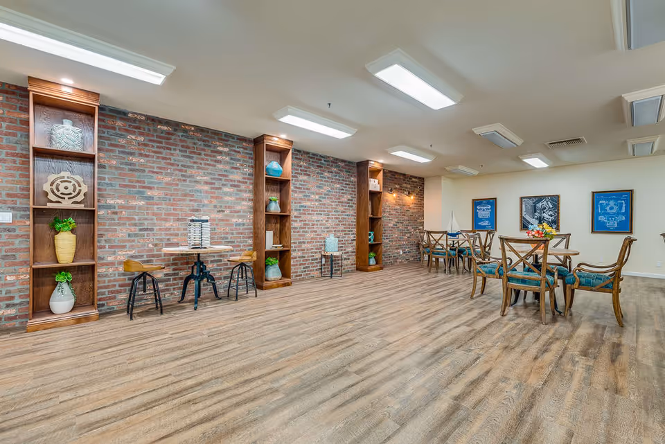 A spacious room with wooden flooring and brick accent walls featuring built-in wooden shelves decorated with vases and ornaments. There are several wooden tables and chairs with teal cushions arranged in the room, along with framed artwork on the far wall. The ceiling has multiple rectangular fluorescent lights.
