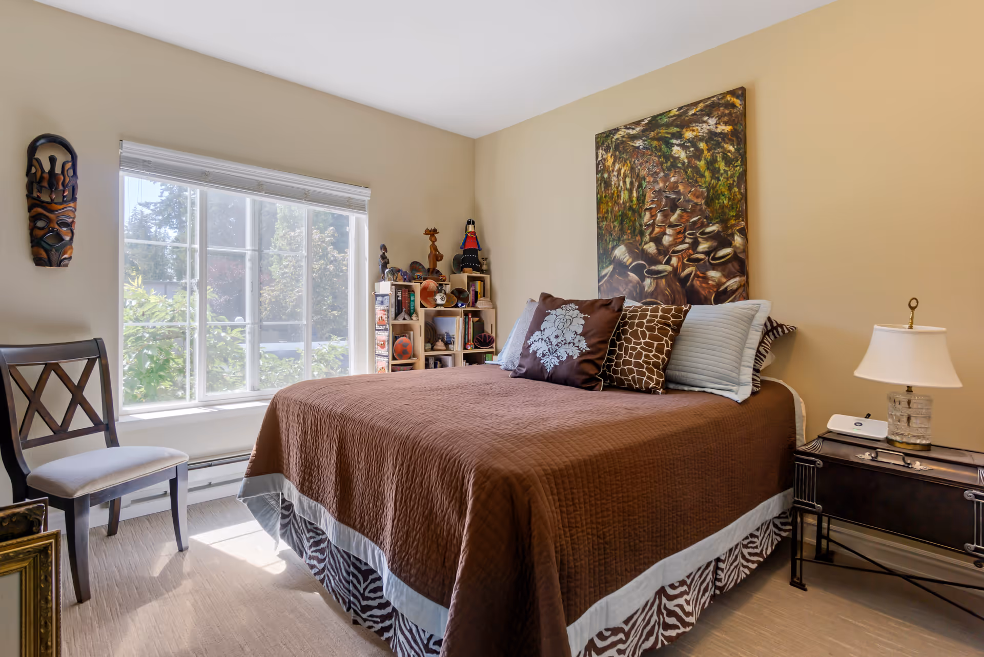 A cozy bedroom with a large window letting in natural light. The bed is covered with a brown quilt and decorated with multiple pillows in various patterns. There is a wooden chair with a cushioned seat near the window, a small nightstand with a lamp, and a colorful painting hanging above the bed. A wooden shelf with decorative items is placed in the corner near the window. The walls are painted beige.