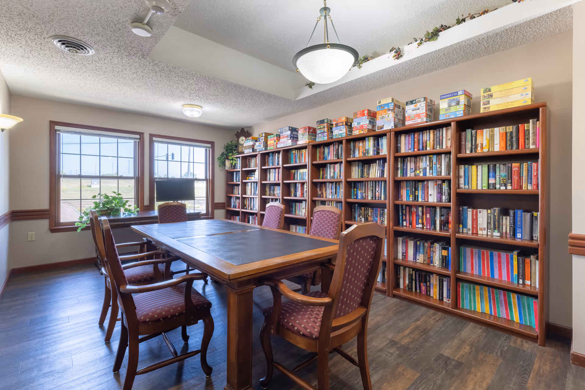 A well-lit room with a large wooden table surrounded by six upholstered chairs. Along one wall are multiple bookshelves filled with books and board games on top. Two large windows let in natural light, and there is a small TV and some plants on the windowsill.