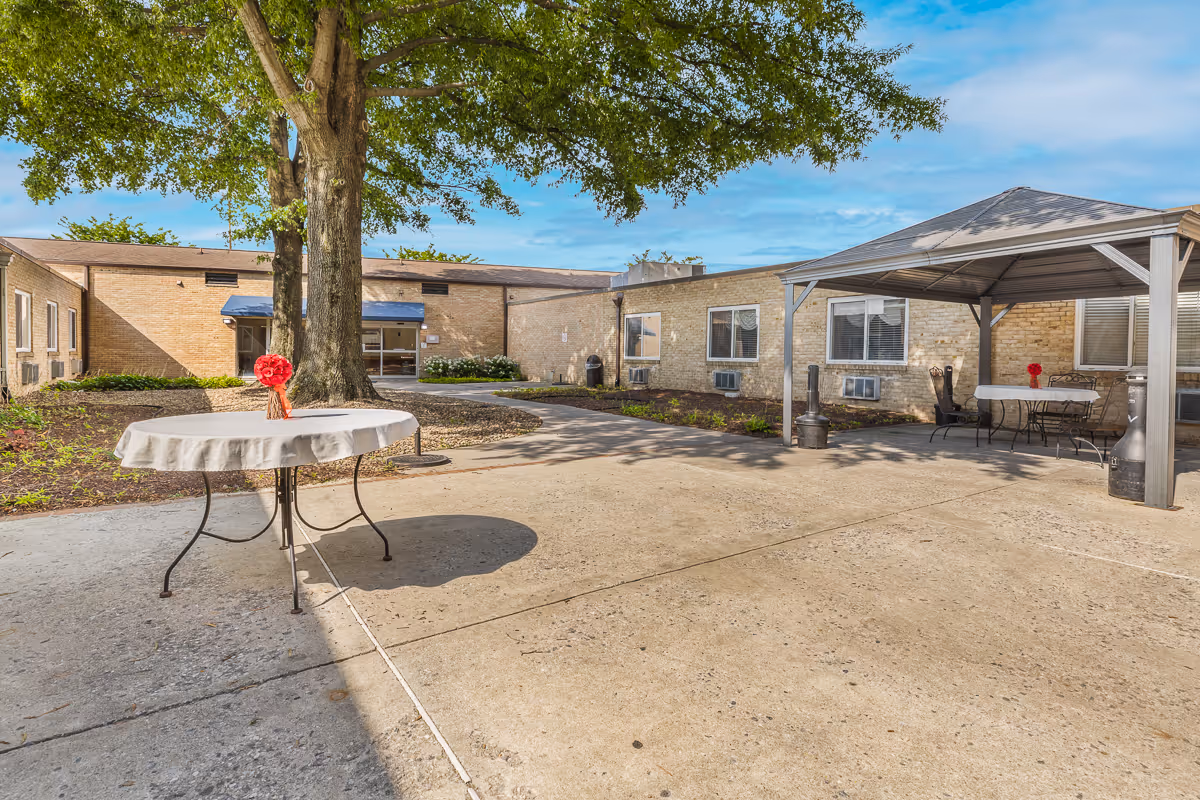 Outdoor courtyard area at Autumn Lake Healthcare at Baltimore Washington featuring a large tree in the center, a round table with a white tablecloth and a red flower centerpiece, a covered seating area with tables and chairs, and a brick building surrounding the courtyard under a blue sky.