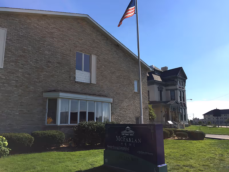 Exterior view of McFarlan Home, a senior living facility, showing a brick building with windows, an American flag on a flagpole, manicured bushes, and a sign in front with the facility's name and contact information. A neighboring older-style house is visible in the background under a clear blue sky.