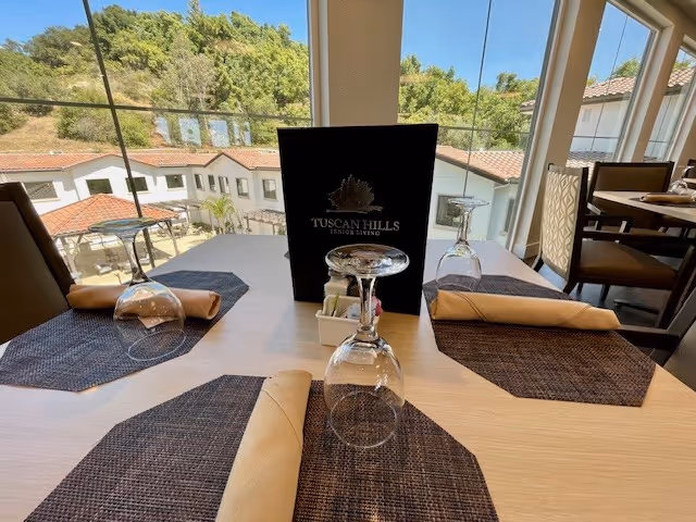 A dining table set for three with beige napkins and upside-down wine glasses on dark placemats, inside a room with large windows showing a view of a hillside and buildings outside. A menu with the Tuscan Hills Senior Living logo is placed in the center of the table.