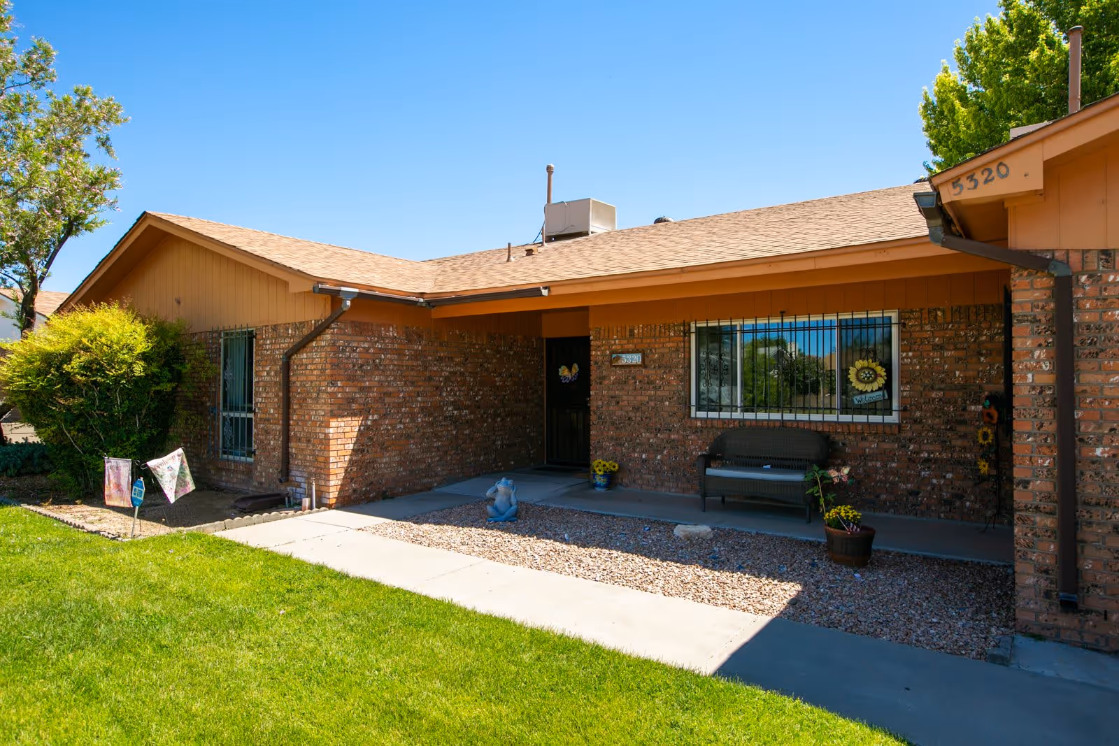 Exterior view of a single-story brick building with a tan roof and a small covered porch area. There is a bench, potted plants, and decorative items near the entrance. The building is surrounded by a well-maintained green lawn and trees under a clear blue sky.