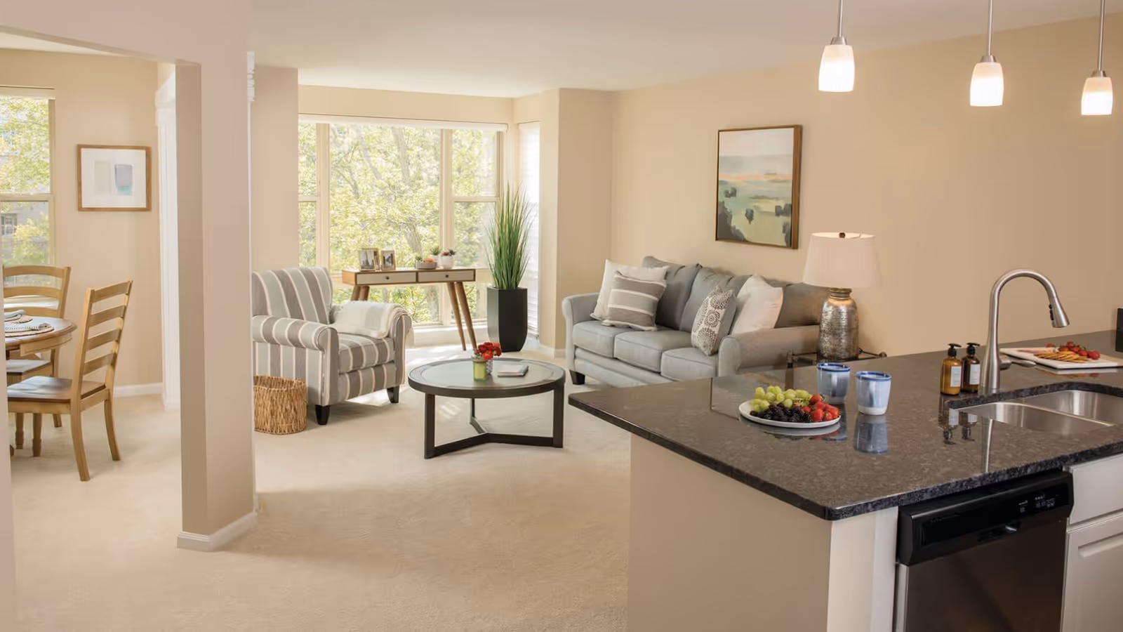 A bright and cozy senior living community interior featuring a living room with a gray sofa, striped armchair, round glass coffee table, and a large window letting in natural light. Adjacent to the living room is a kitchen area with a black granite countertop, stainless steel sink, and modern fixtures. In the background, a dining area with a wooden table and chairs is partially visible.