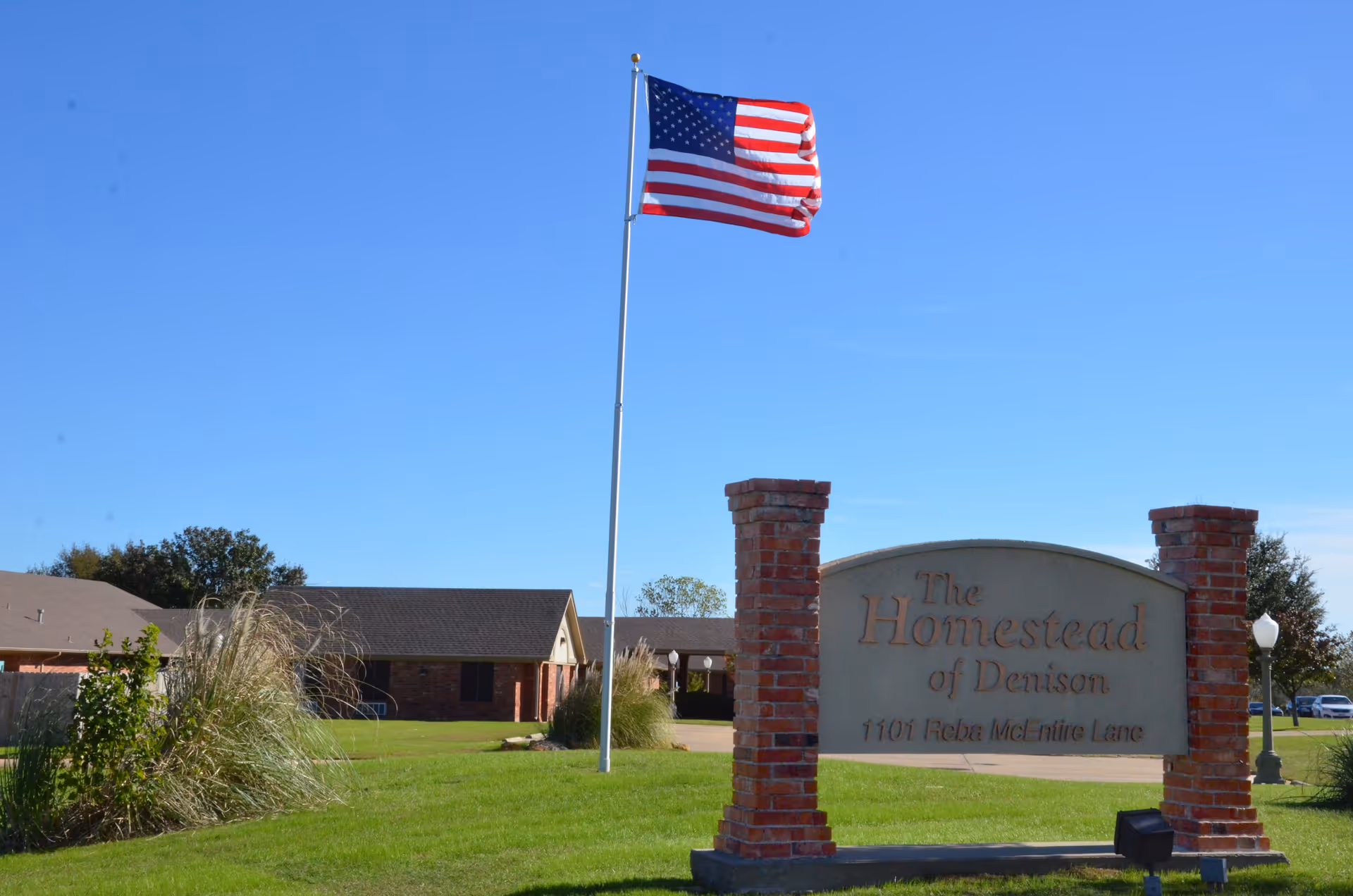 Outdoor view of The Homestead of Denison facility sign with an American flag flying on a flagpole against a clear blue sky. The building and green lawn with some plants are visible in the background.
