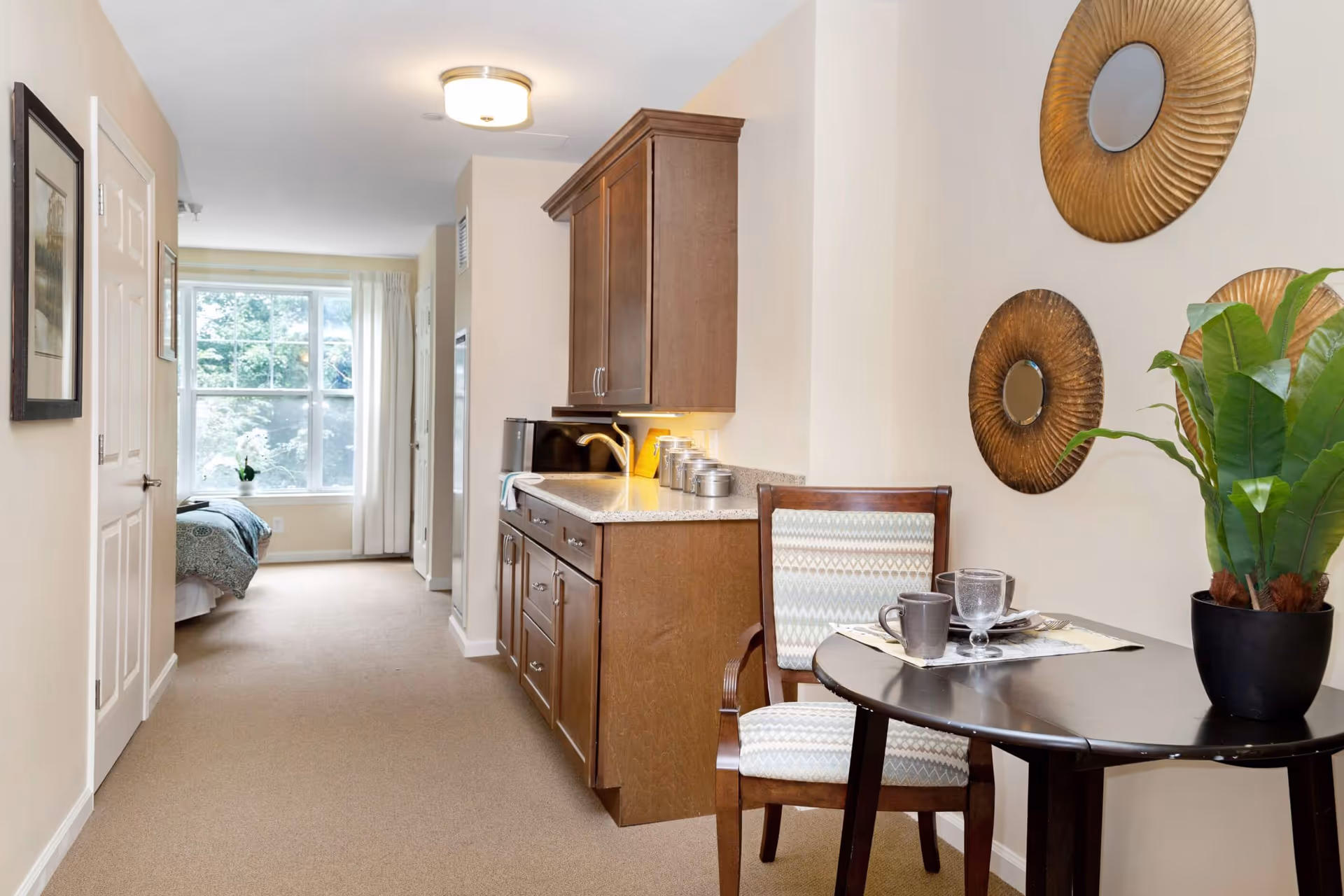 Interior view of a senior living facility room showing a small dining area with a round table, a chair, and a potted plant. On the wall above the table are three decorative round mirrors. To the left is a kitchenette with wooden cabinets and a countertop with kitchen items. In the background, a bedroom area with a bed and a large window with white curtains is visible.