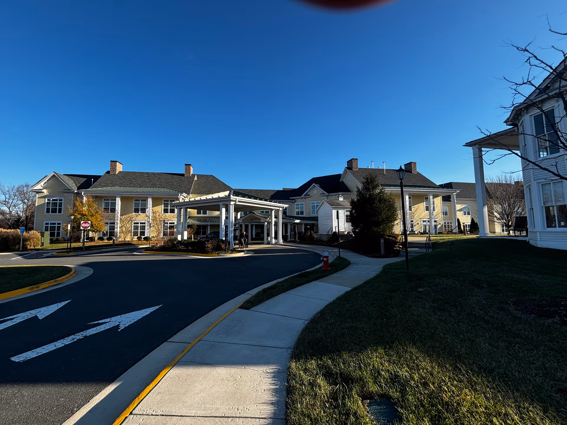 Exterior view of Sunrise at Silas Burke House, a large yellow senior living facility with white trim and multiple chimneys. The building has a covered entrance with a driveway and a sidewalk leading up to it. The sky is clear and blue, and there is well-maintained grass and some trees around the building.