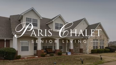 Exterior view of a single-story senior living facility building with a combination of siding and stone facade, multiple windows, and a front porch with seating. The sky is overcast.