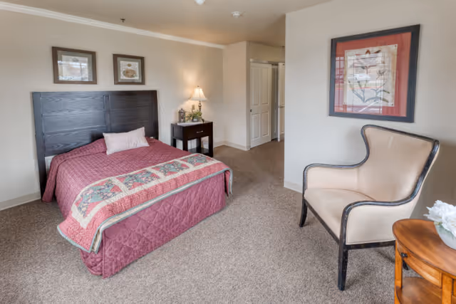 A senior living bedroom with a bed covered in a maroon quilt and a decorative throw. There is a dark wooden headboard, two framed pictures above the bed, a nightstand with a lamp and small plant, a beige upholstered armchair, and a framed floral artwork on the wall. The room has beige walls and carpeted flooring.