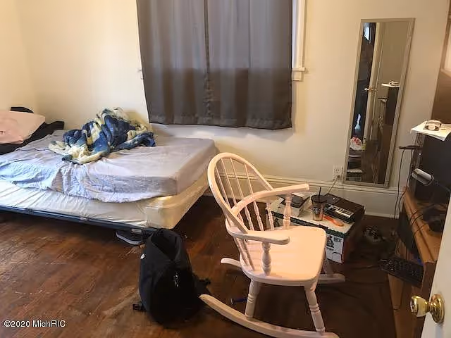 A small bedroom with a mattress on a metal frame against the wall, covered with a blue and white blanket and a pink pillow. A wooden rocking chair is positioned near the mattress. There is a black backpack on the wooden floor next to the mattress. A full-length mirror is mounted on the wall near a small table holding various items including a drink cup and electronic devices. A window with closed gray curtains is above the mattress.