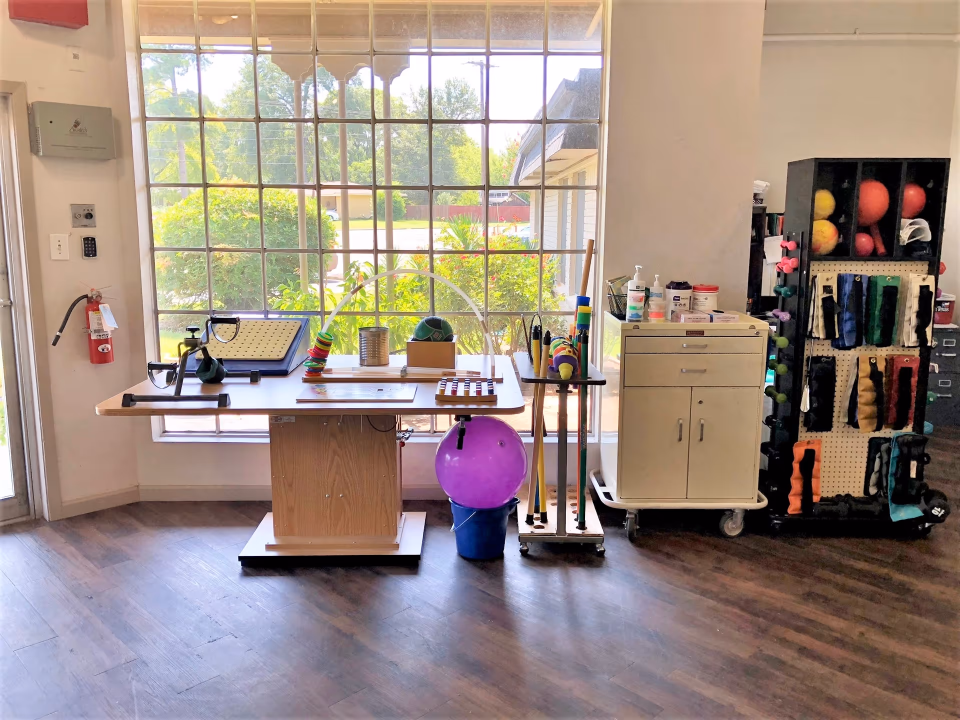 A therapy or activity room with a large window letting in natural light. In front of the window is a wooden table with various therapy tools and equipment, including a purple exercise ball, colorful rings on a rod, and other small items. To the right, there is a white rolling cabinet with hand sanitizer and other supplies on top, and a black shelving unit holding various exercise balls and weights. The floor is wooden, and the room appears clean and organized.
