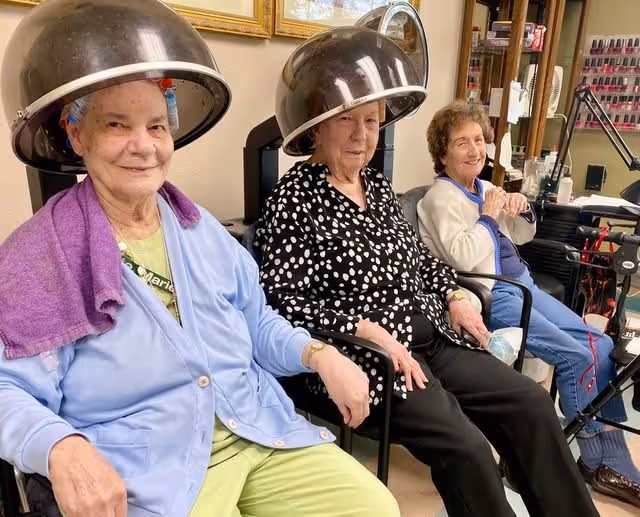 Three elderly women sitting side by side in a salon area, two of them under hair drying machines, smiling and relaxing.