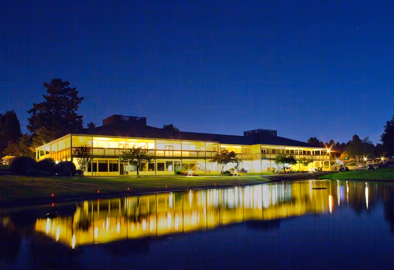 Nighttime view of a large two-story building with many windows illuminated from inside, reflecting on a calm body of water in front of it, surrounded by trees and a well-maintained lawn.