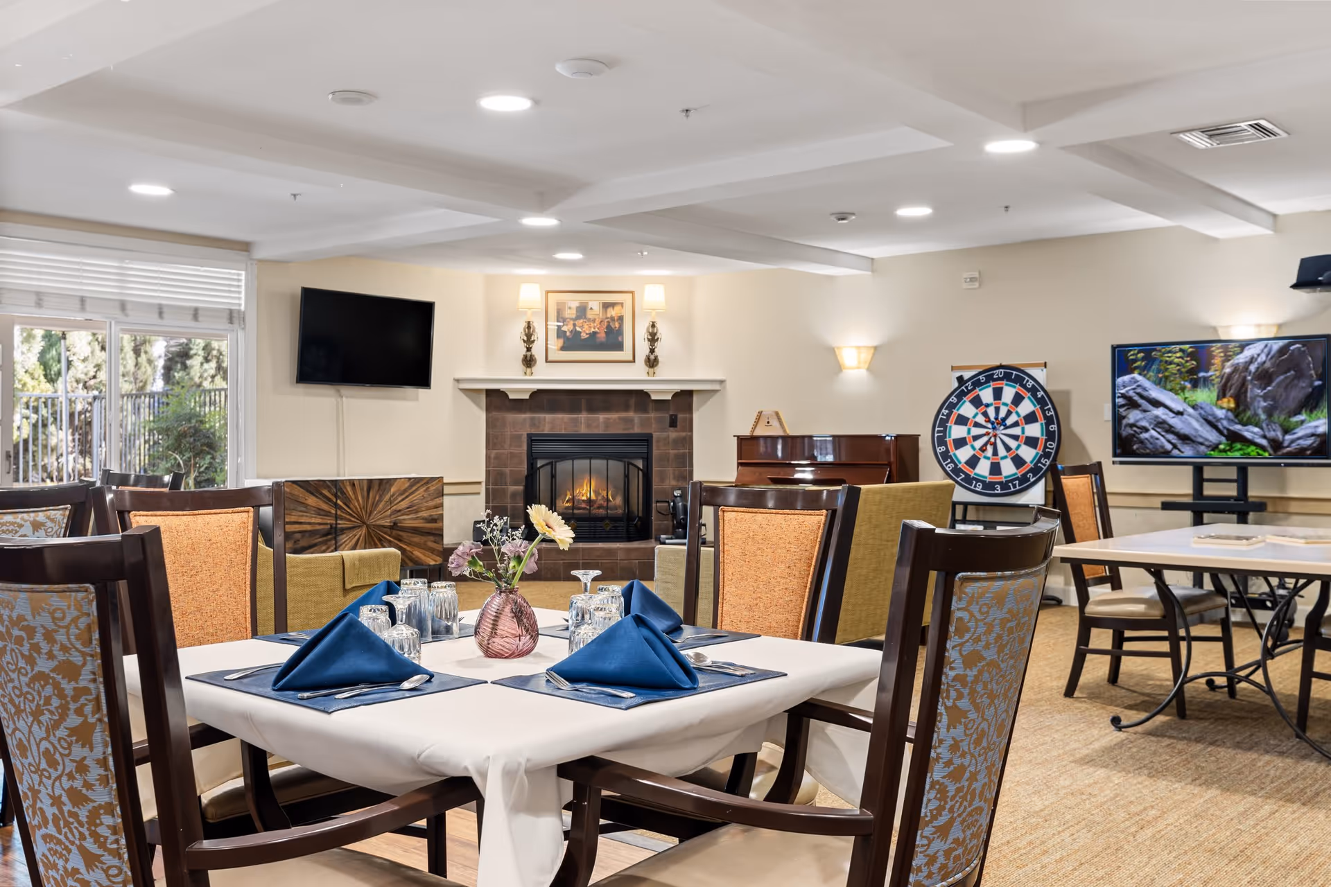 A well-lit common area with dining tables set with blue napkins, glasses, and a small flower vase. The room features a fireplace with a framed picture and two wall sconces above it, a flat-screen TV mounted on the wall, a dartboard, a piano, and another TV displaying a nature scene. Large windows with blinds allow natural light to enter the room.