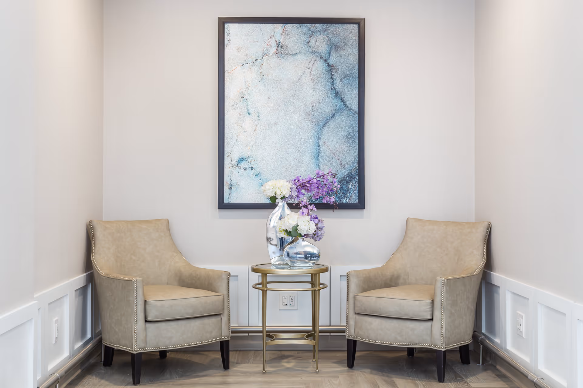 Two beige armchairs with nailhead trim facing each other with a small round gold table between them holding two glass vases with white and purple flowers. A large abstract blue and white painting is hung on the wall behind the table. The room has light-colored walls and wainscoting with a wood floor.