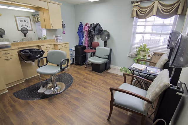 A small salon room with a styling chair and sink, waiting chairs, hairdryers and a window with a valance.