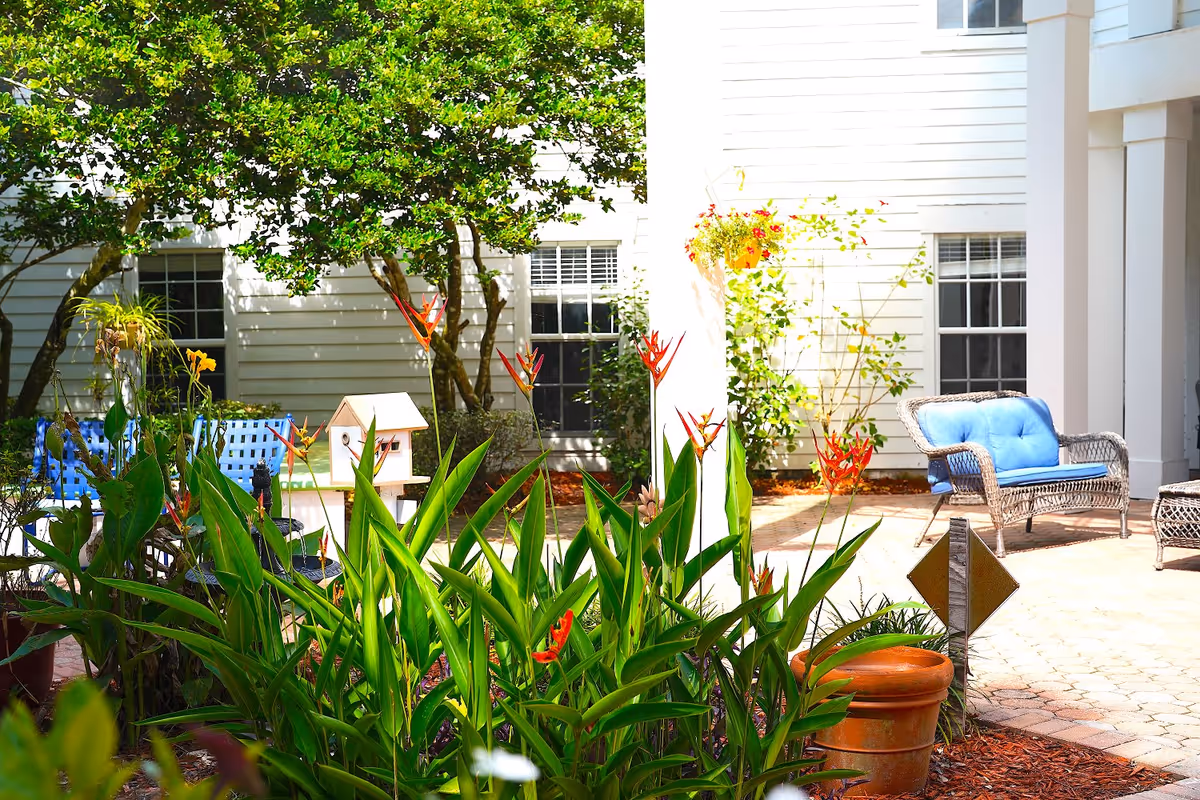 Outdoor garden area with green plants and red flowers in the foreground, a white birdhouse, and wicker patio furniture with blue cushions on a paved patio. The background shows white siding walls with windows and some trees providing shade.