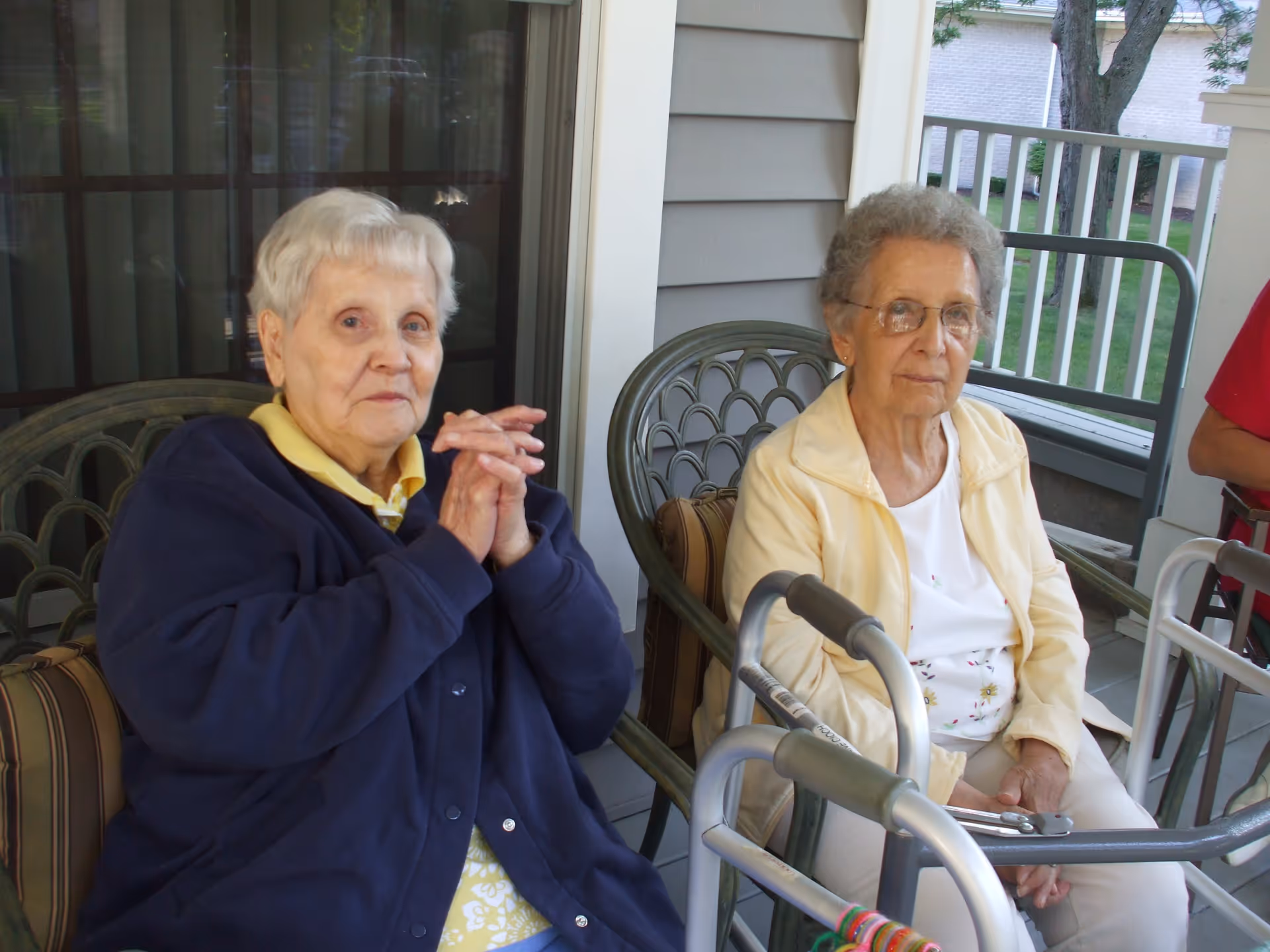 Two elderly women sitting on a porch or patio area with chairs. One woman is wearing a dark blue jacket and yellow shirt, and the other is wearing a light yellow jacket and glasses. A walker is positioned in front of the woman in the light yellow jacket. The background shows a railing and some greenery outside.