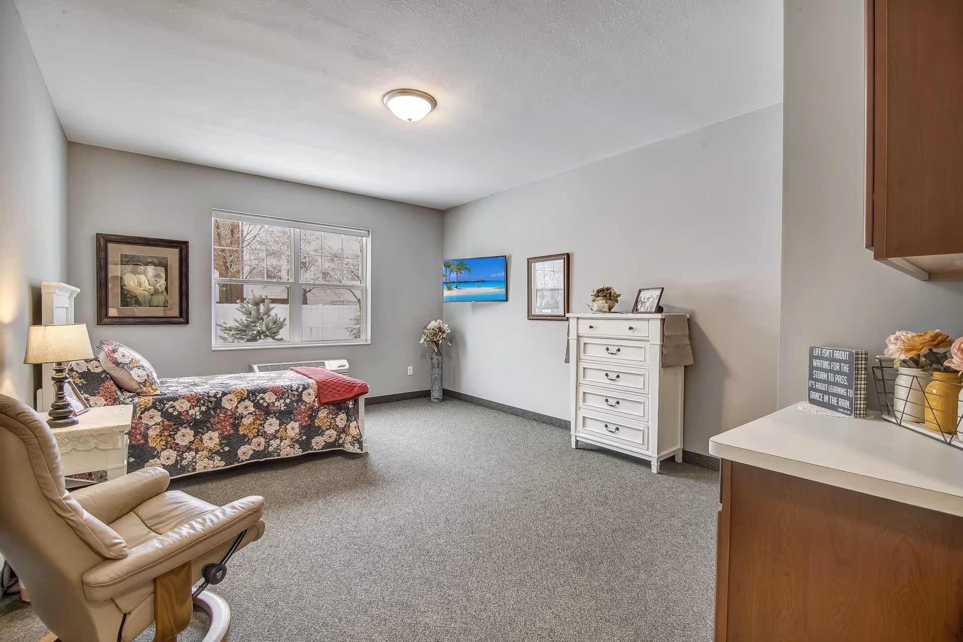 A furnished bedroom with a single floral bed, recliner, white dresser, and wall-mounted TV in a senior living facility.