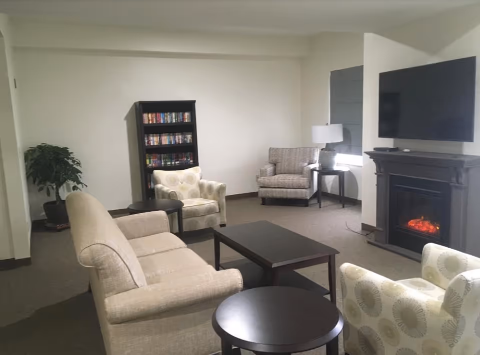 A cozy living room with beige upholstered sofa and armchairs arranged around a dark wooden coffee table. There is a black bookshelf filled with books against the wall, a potted plant in the corner, and a fireplace with a mounted flat-screen TV above it. A side table with a lamp is positioned next to one of the armchairs.