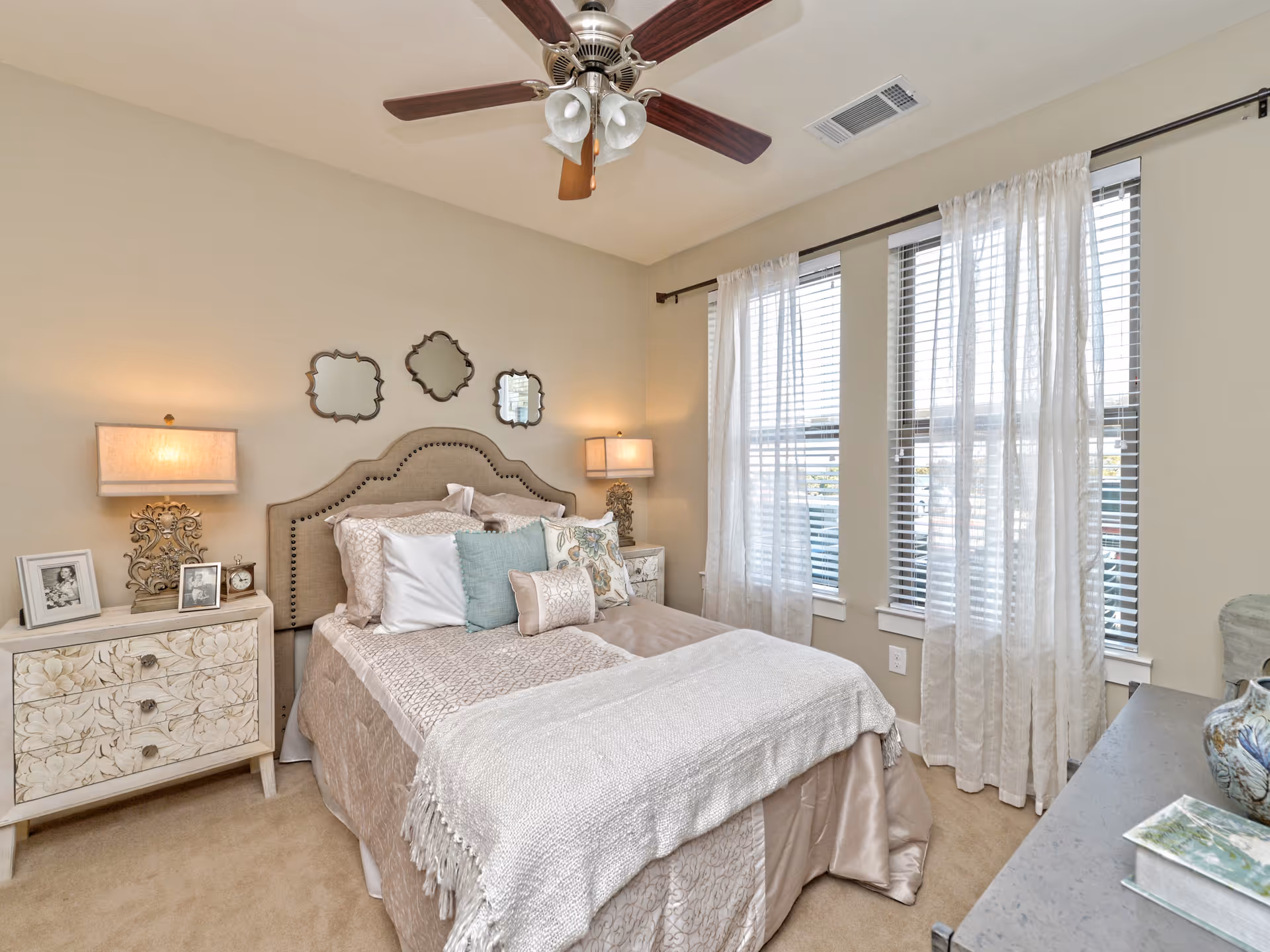 A cozy bedroom with a beige upholstered bed adorned with multiple pillows and a light beige comforter. There are two matching bedside tables with decorative lamps and framed photos. Three decorative mirrors hang above the headboard. The room has beige walls, a ceiling fan with wooden blades, and three tall windows with white blinds and sheer curtains, allowing natural light to fill the space.