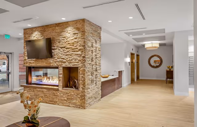 Interior view of a senior living facility lobby featuring a modern stone fireplace with a flat-screen TV mounted above it, light wood flooring, and a hallway leading to other rooms. There is a round table with a flower arrangement in the foreground and a decorative mirror on the far wall.