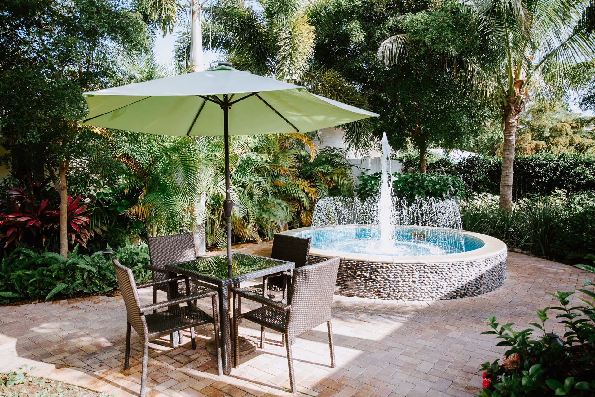 Outdoor patio area with a round stone fountain spraying water, surrounded by lush green palm trees and plants. There is a square glass-top table with four wicker chairs and a large green umbrella providing shade.