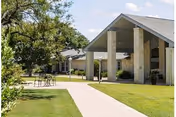 Outdoor view of a senior living facility with a paved walkway leading to a covered entrance supported by large stone pillars. There is a small round table with chairs on the grass near the walkway, and trees and well-maintained lawns surround the area under a partly cloudy sky.