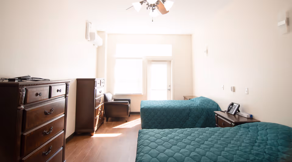 A bright bedroom with two twin beds covered in teal quilted bedspreads. The room features wooden dressers, a dark armchair near a window with blinds, and a door leading outside. The walls are painted white, and there is a ceiling fan with lights.