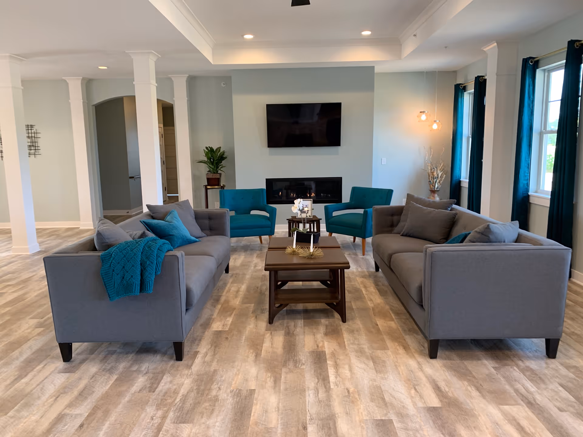 Spacious common living room with two gray sofas, teal armchairs, a coffee table, wall-mounted TV and fireplace near large windows.
