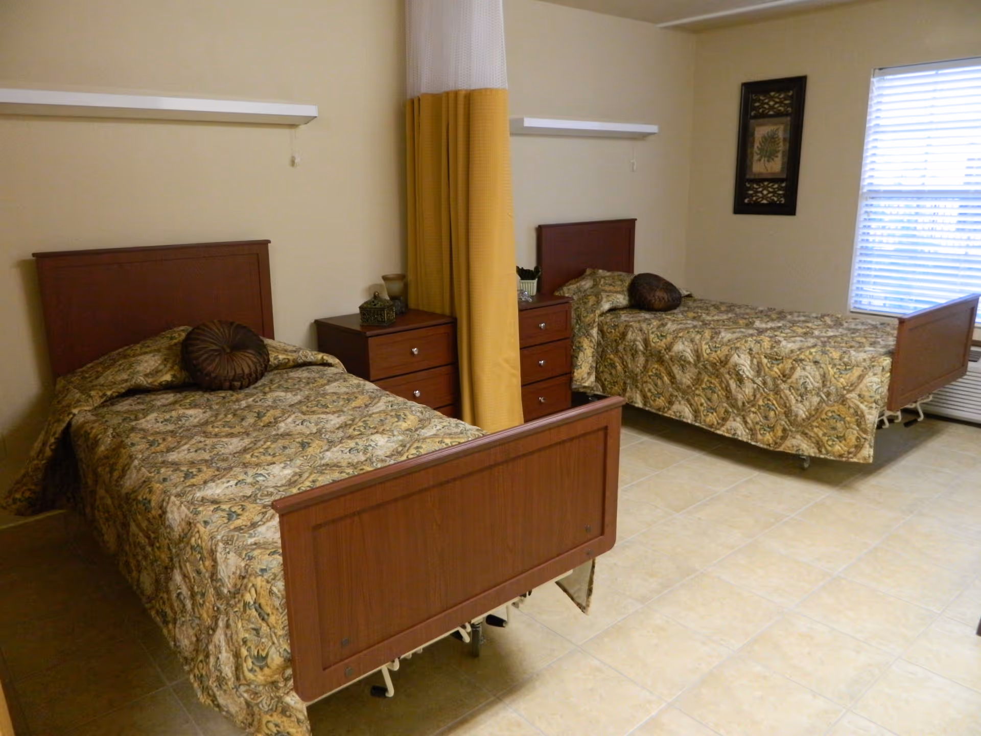 A room with two single beds separated by a yellow and white privacy curtain. Each bed has a patterned bedspread and a round decorative pillow. There are two wooden nightstands with drawers between the beds, and a window with blinds on the right wall. The floor is tiled and the walls are painted beige.