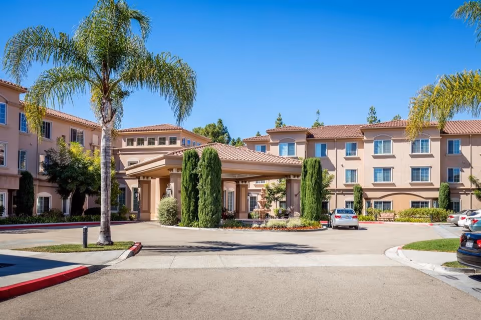 Exterior view of Fairwinds - Ivey Ranch senior living facility showing a three-story building with beige walls and red-tiled roof, surrounded by palm trees and greenery under a clear blue sky. Several cars are parked near the entrance driveway.