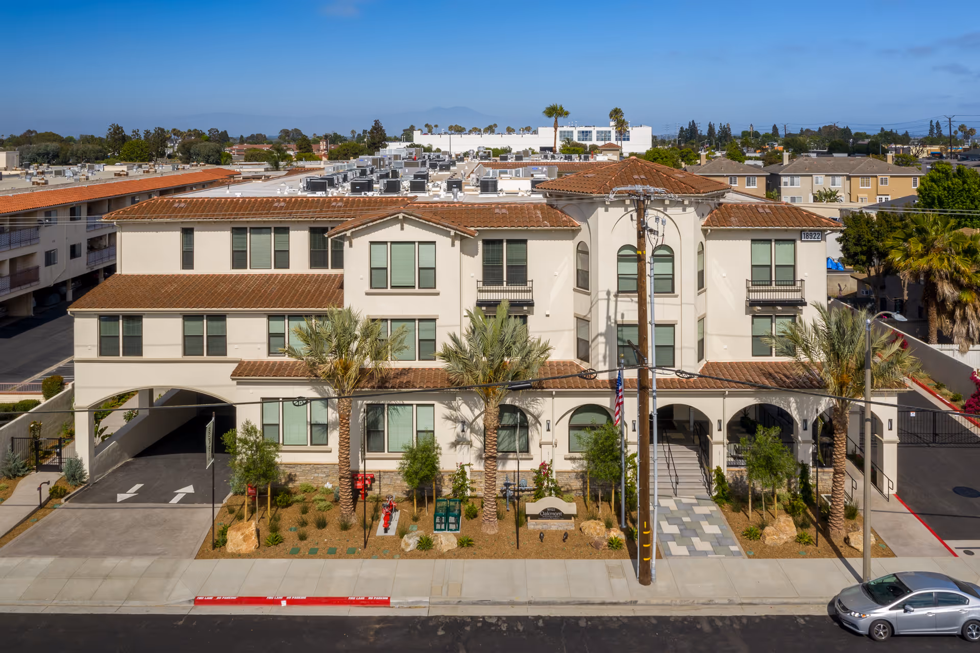 Front exterior view of Oakmont of Huntington Beach, a three-story building with a beige facade, red tile roof, and multiple windows. The entrance features arches, palm trees, and a landscaped area with shrubs and rocks. A car is parked on the street in front, and the sky is clear and blue.